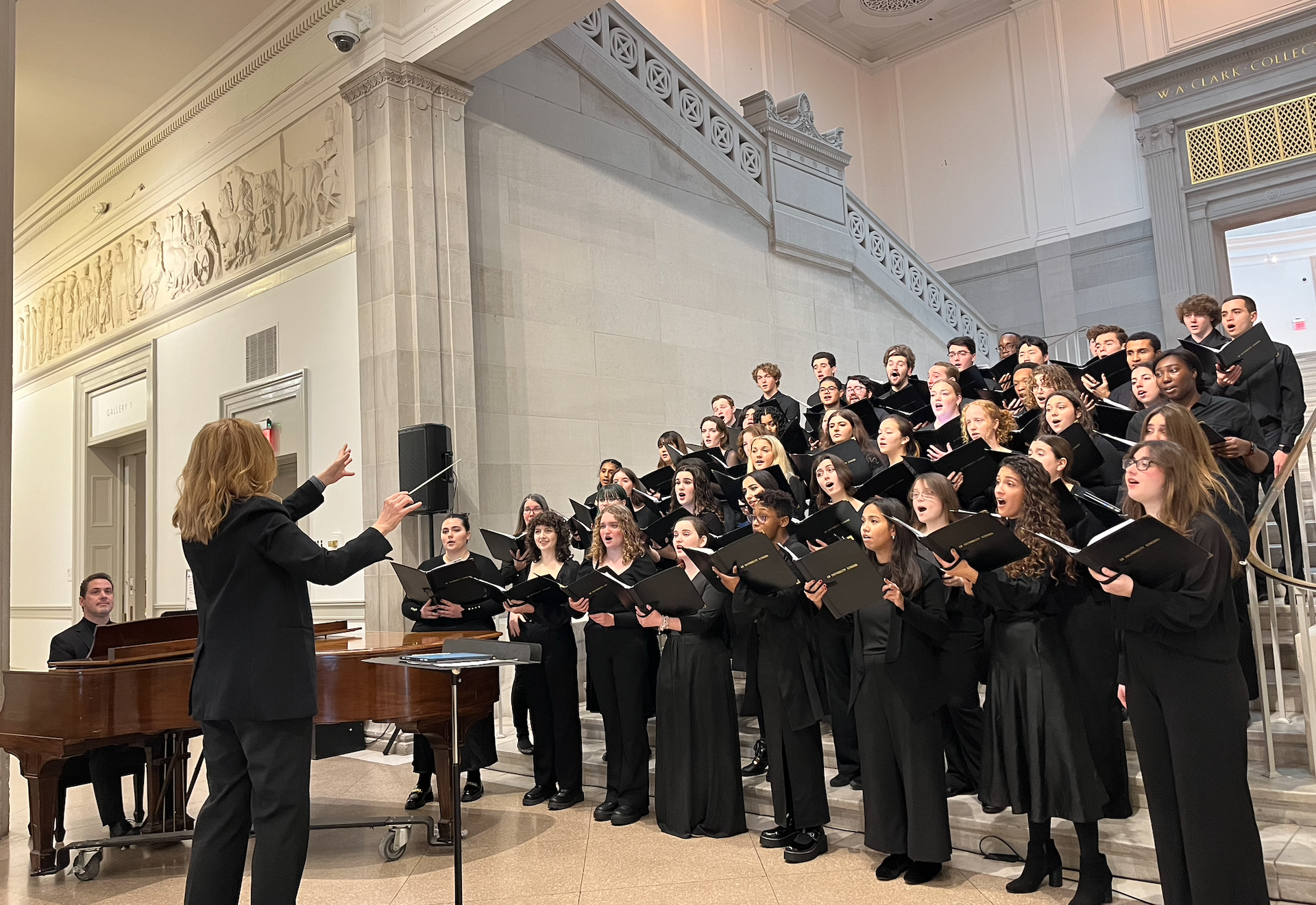 Conductor Erin Freeman directing the University Singers, who are dressed in black concert attire, standing in rows on steps. Next to Erin Freeman (also dressed in black) is a man seated at and playing a brown grand piano.