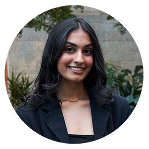 a woman with dark hair and a black blazer in front of a stone wall with plants 