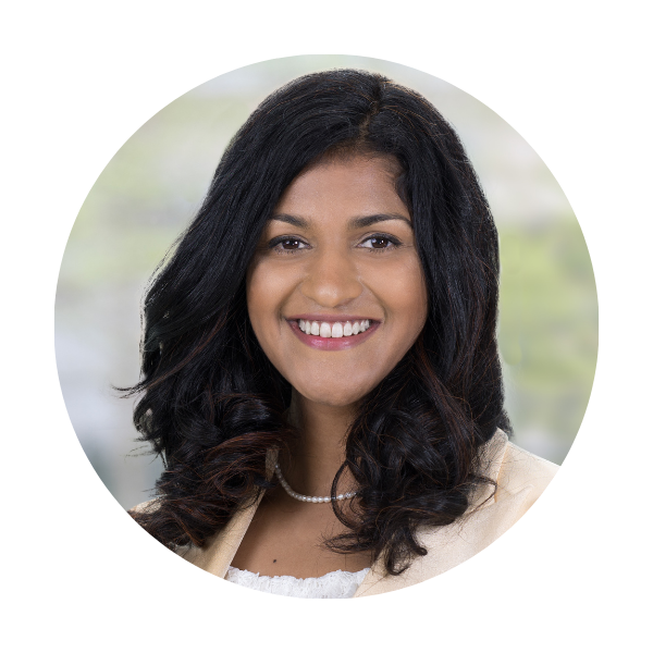 a woman with long dark hair and a necklace and a cream blazer in front of a warm green outdoor background