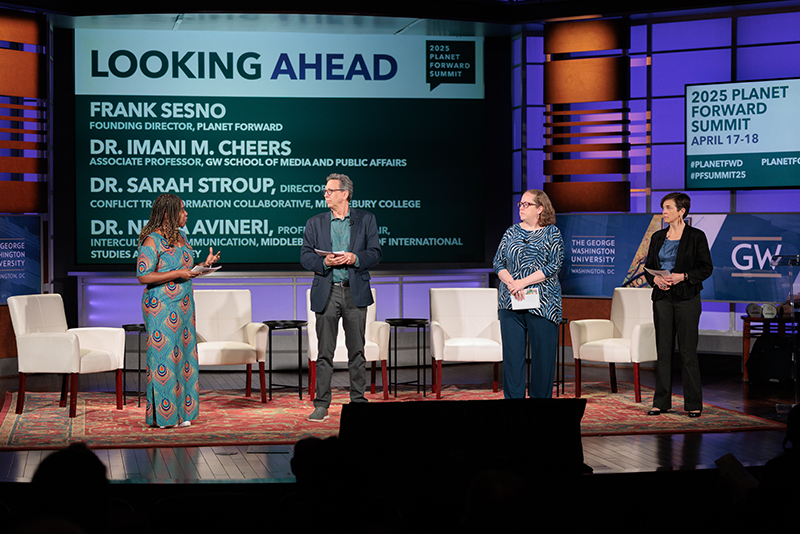 Four people on stage with white chairs behind them, a woman in a colorful dress, a man in a navy sport coat, a woman in a blue shirt, and a woman in a black suit
