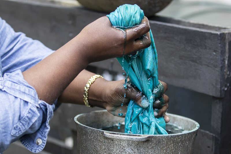 Graduation Cord Dying Two hands with a gold bracelet squeeze blue dye from a piece of fabric over a bucket