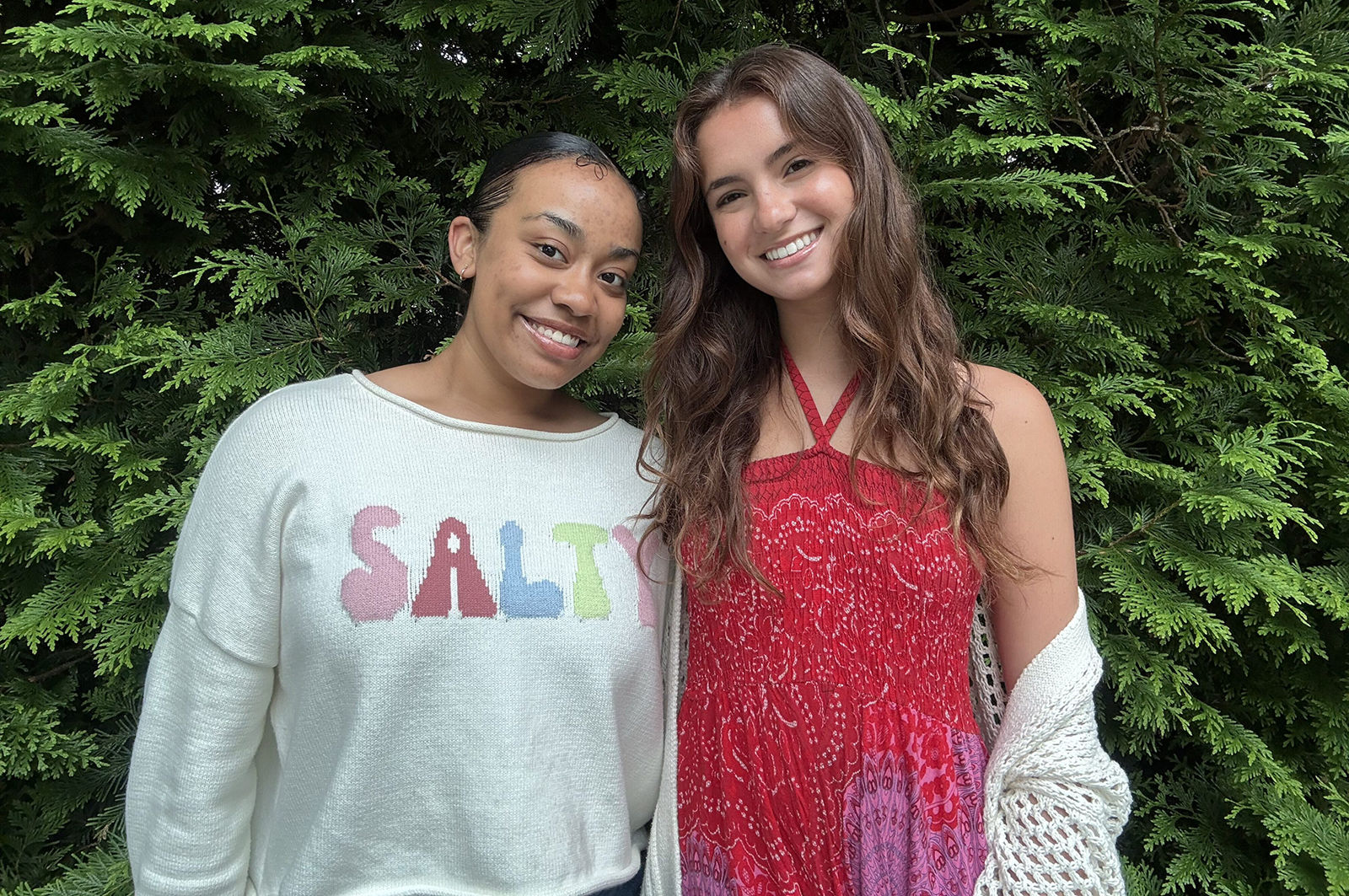 two people stand in front of a green bush one in a grey sweatshirt and the other in a red tank top