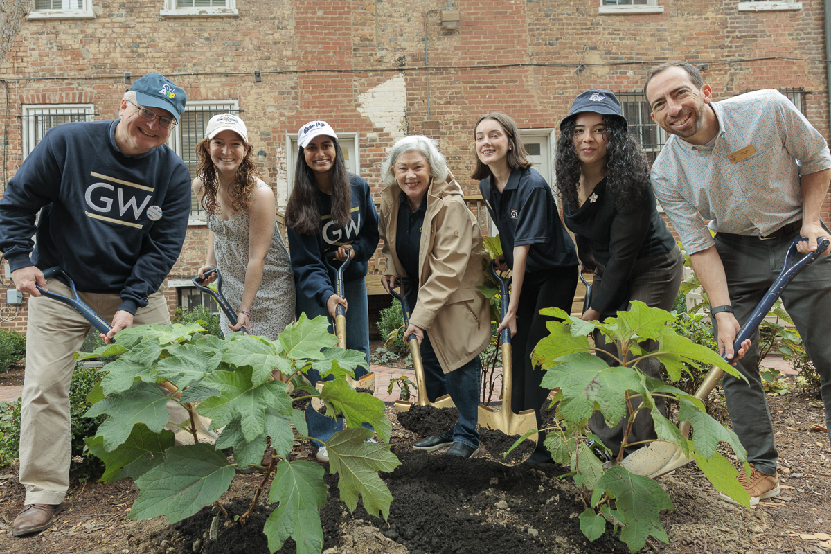 A group of seven people stand in GW buff and blue sweatshirts and hold shovels digging into plants and a garden