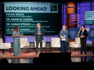 Four people on stage with white chairs behind them, a woman in a colorful dress, a man in a navy sport coat, a woman in a blue shirt, and a woman in a black suit