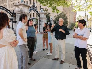 A group of seven students talk with a professor in a black shirt on a sunny day outside of a gate on a college campus.