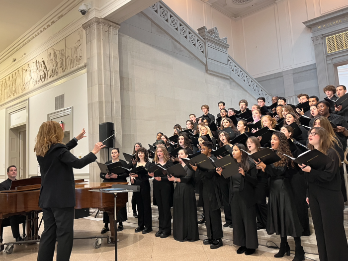Conductor Erin Freeman directing the University Singers, who are dressed in black concert attire, standing in rows on steps. Next to Erin Freeman (also dressed in black) is a man seated at and playing a brown grand piano.