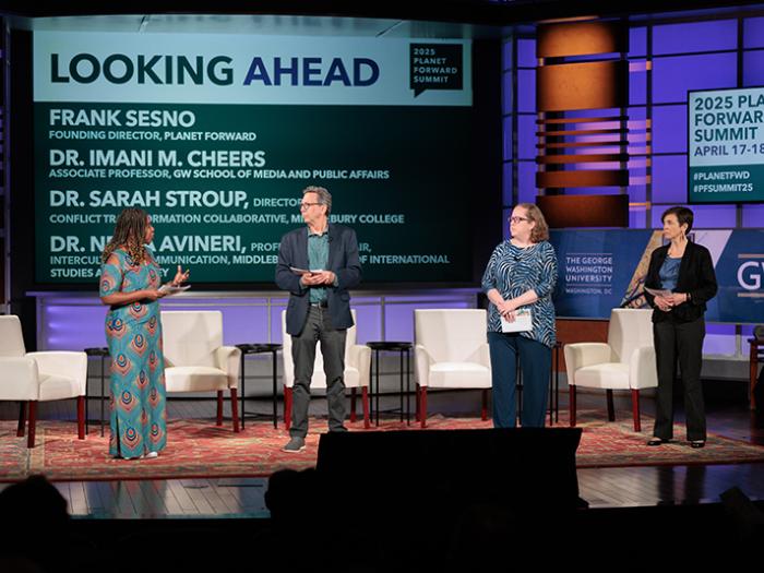 Four people on stage with white chairs behind them, a woman in a colorful dress, a man in a navy sport coat, a woman in a blue shirt, and a woman in a black suit