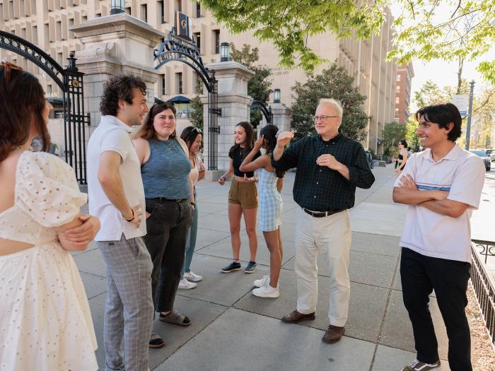 A group of seven students talk with a professor in a black shirt on a sunny day outside of a gate on a college campus.