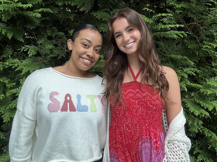 two people stand in front of a green bush one in a grey sweatshirt and the other in a red tank top