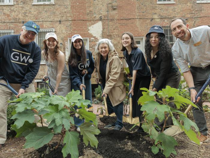 A group of seven people stand in GW buff and blue sweatshirts and hold shovels digging into plants and a garden