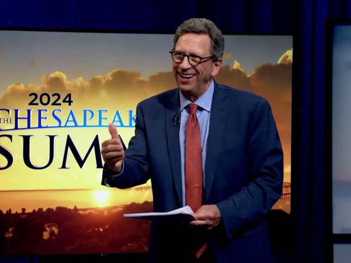 a man in a blue suit and red tie in front of a screen with a sunset with text that reads: "2024 Chesapeake Bay Summit"
