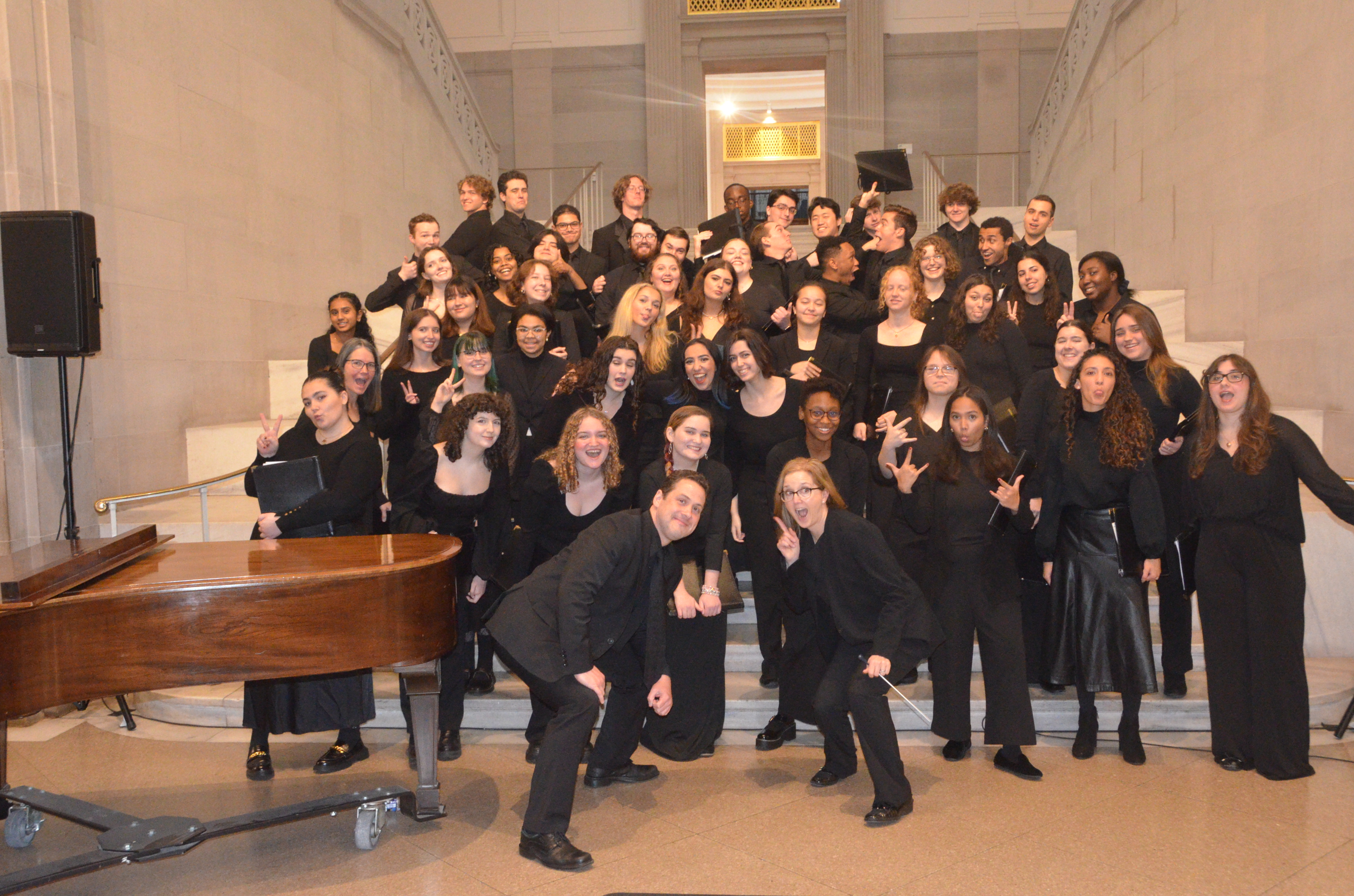 A silly group photo of the University Singers, dressed in black concert, and standing in rows behind a piano.