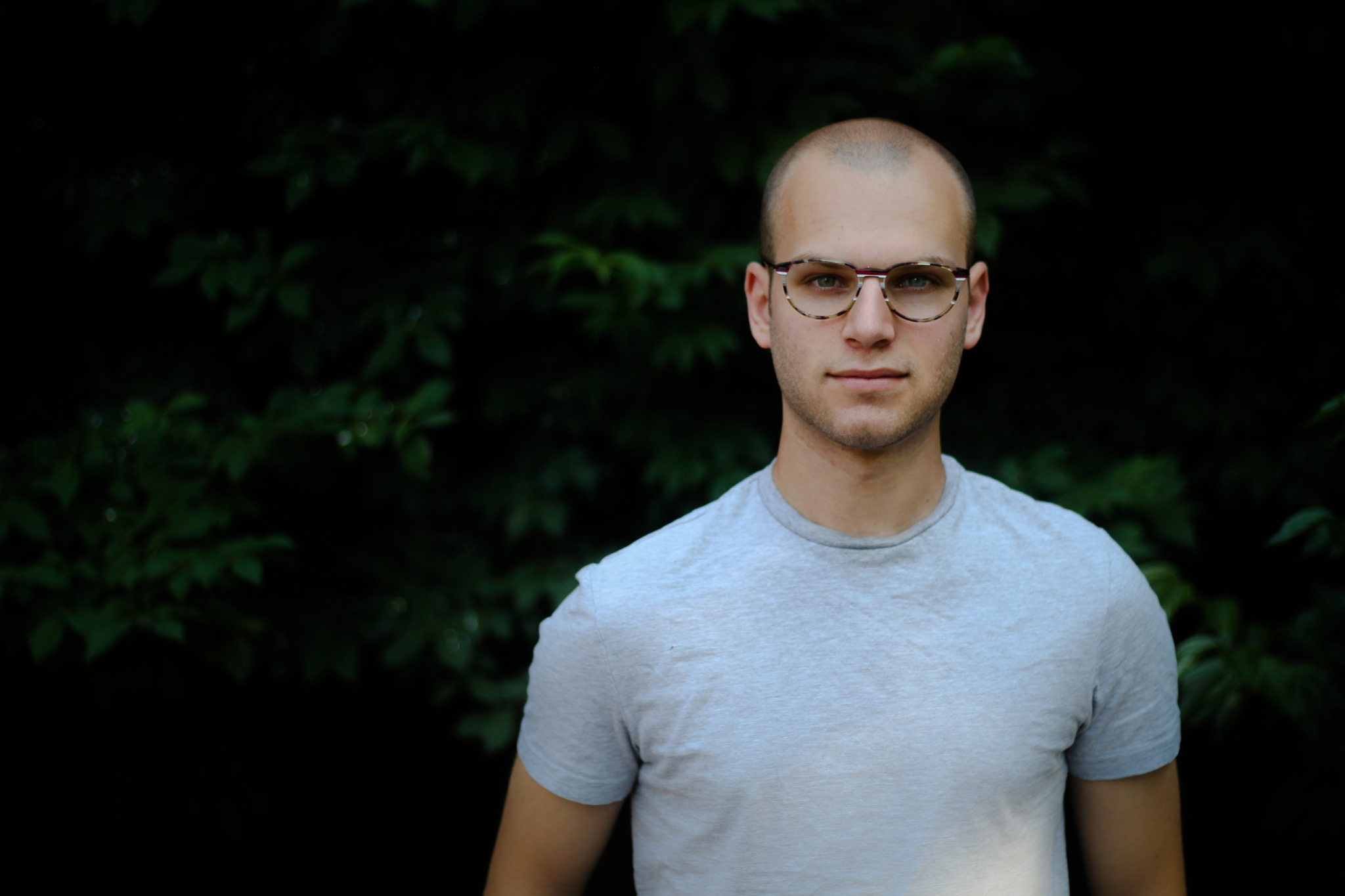 A photo of the composer Alex Berko, wearing a white tshirt and glasses, standing in front of a leafy background.