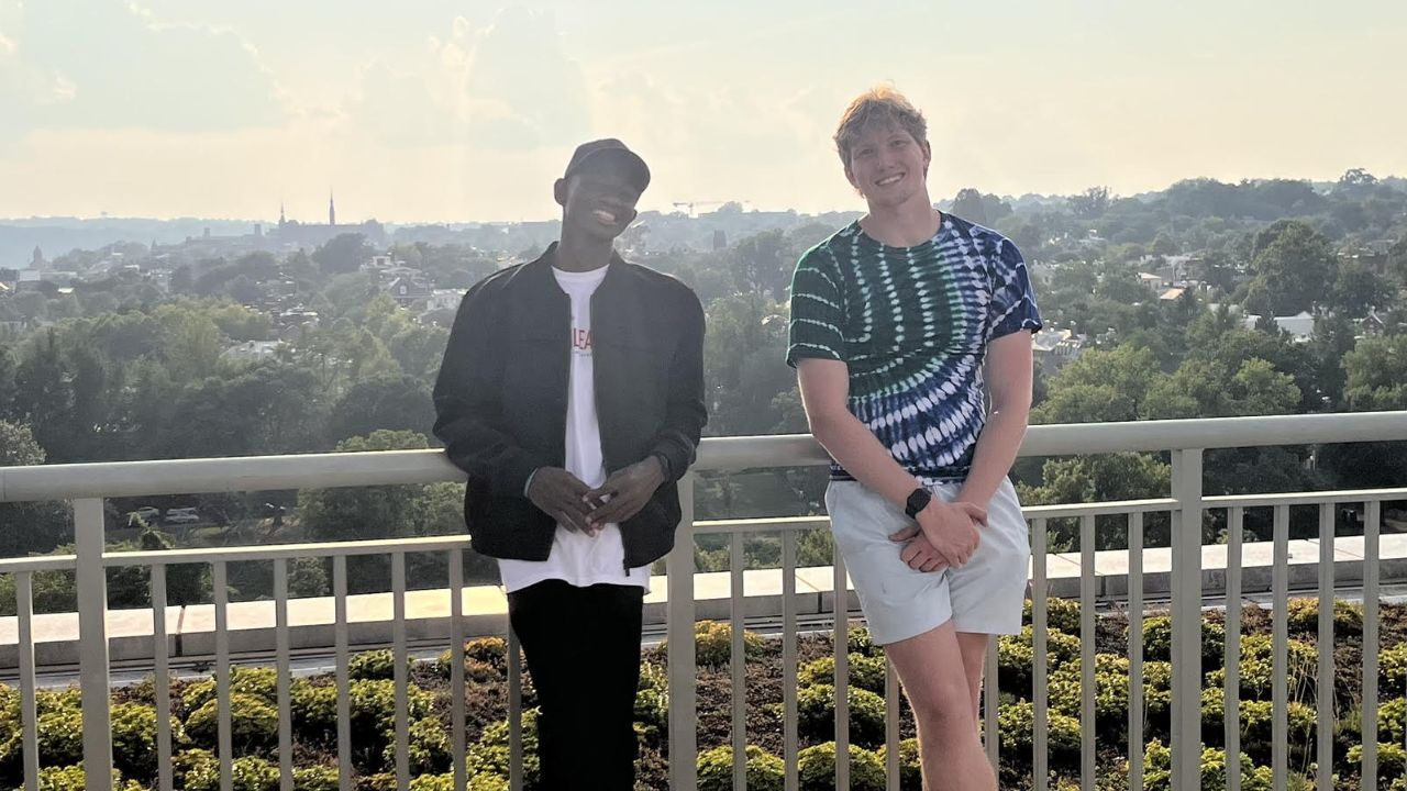 two young men standing on a railing one in a black jacket and one in a tie-dye shirt
