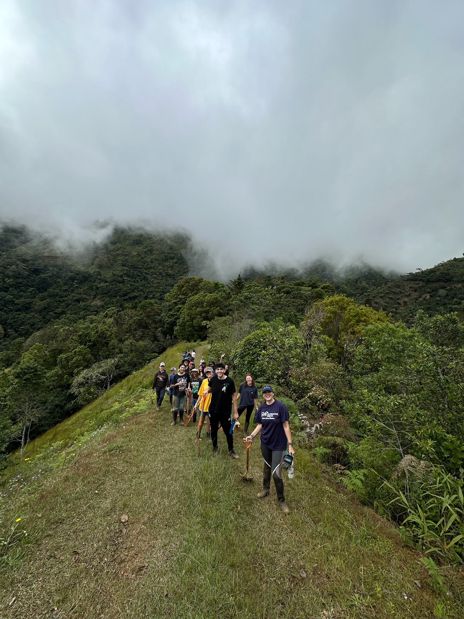 GW Alternative breaks cohort on a hike with shovels, the group is posed in a single-file line on a hill clearing in the jungle, low clouds are overhead.