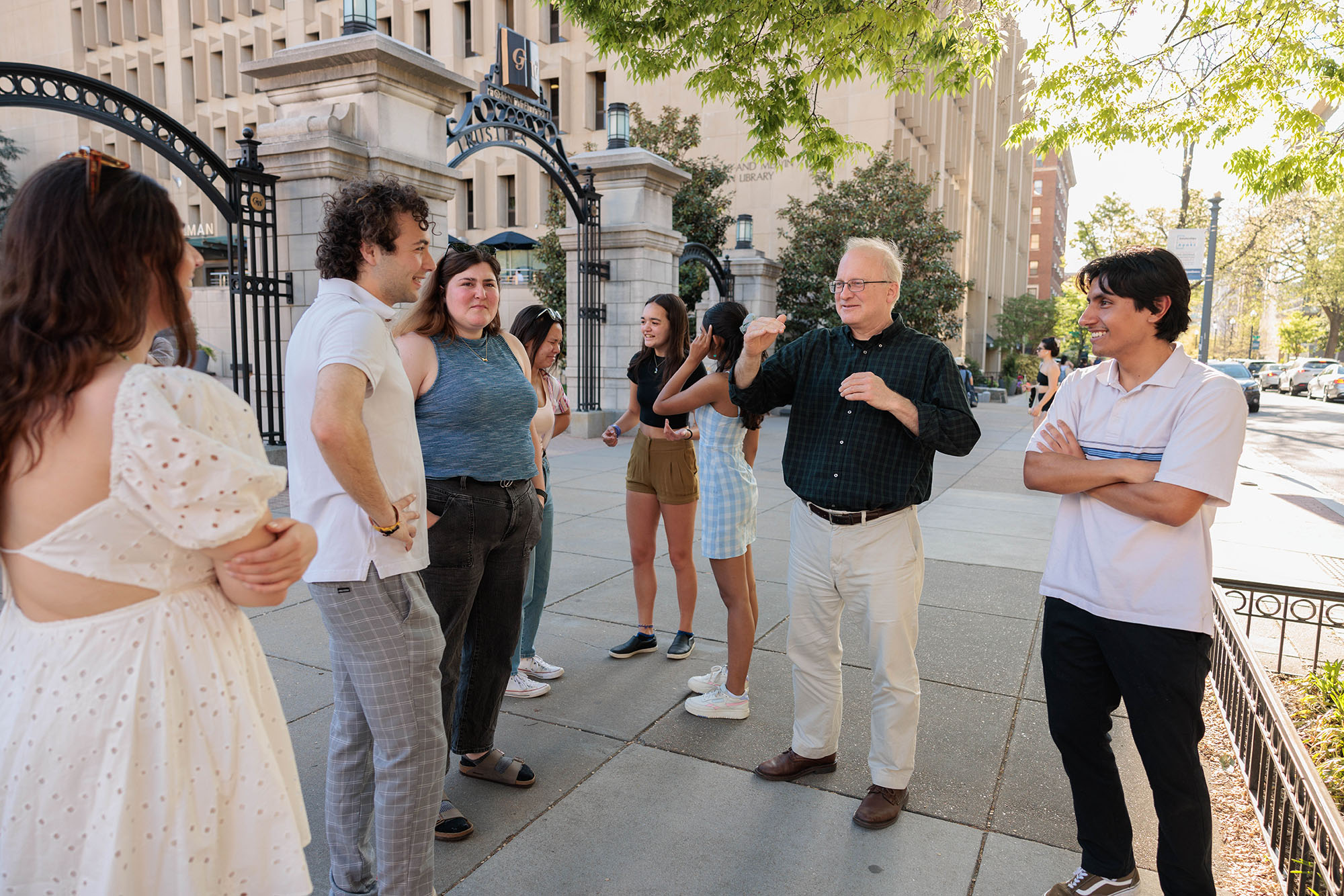A group of seven students talk with a professor in a black shirt on a sunny day outside of a gate on a college campus.
