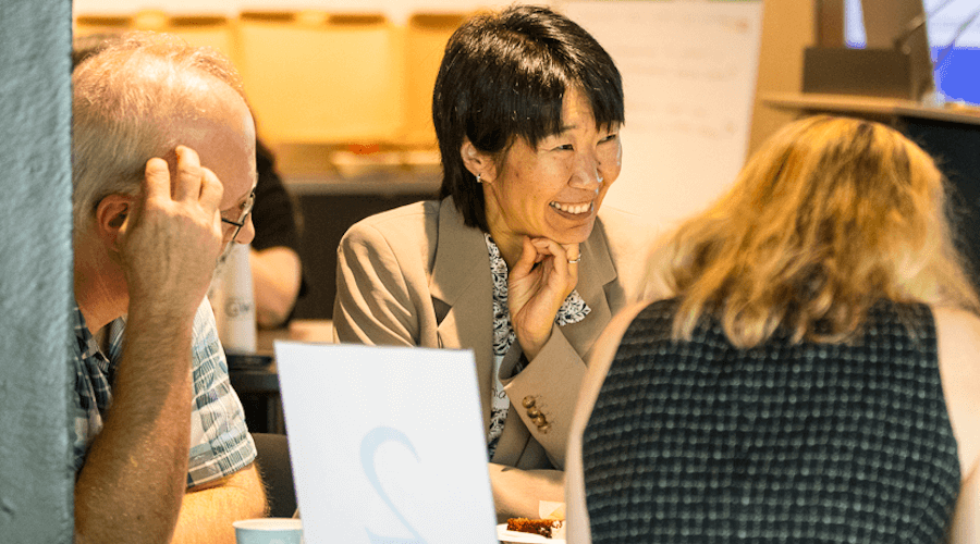 a woman with short hair and a beige blazer speaks to people at a table