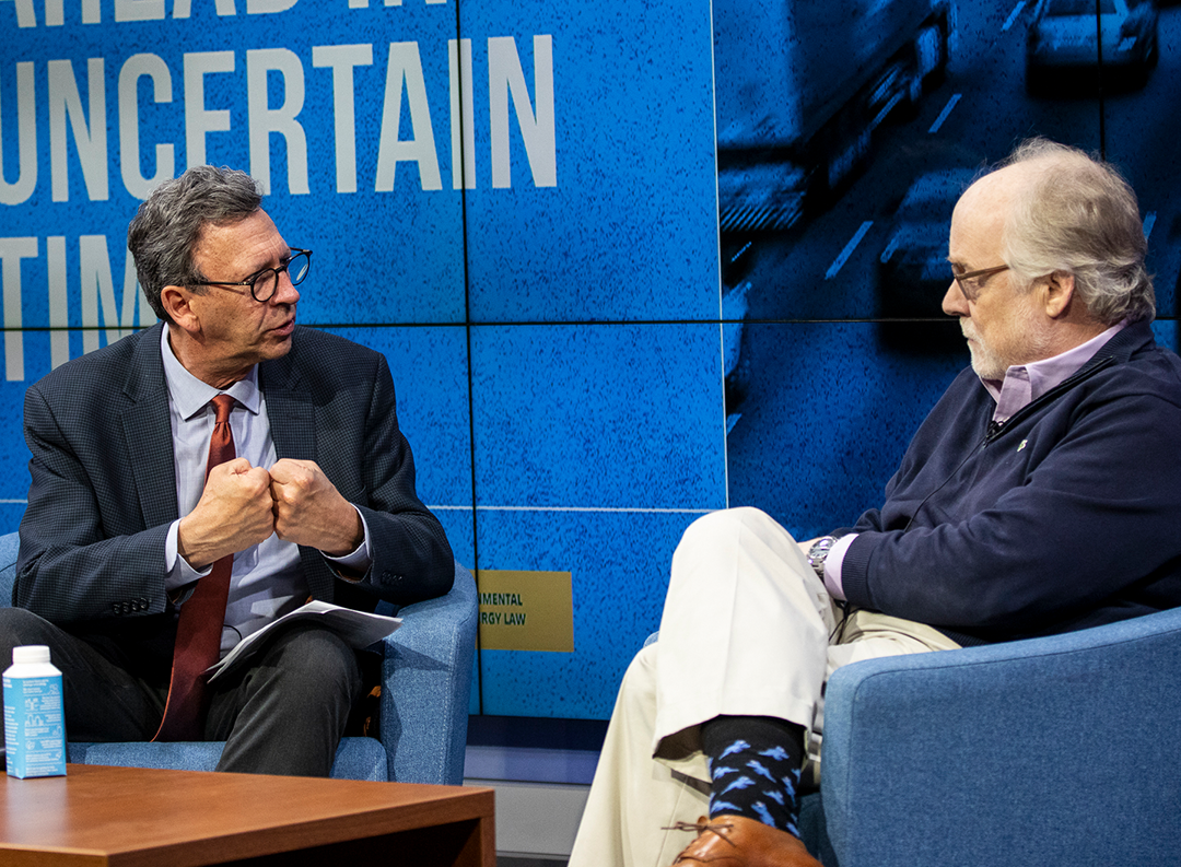 two men one in a suit and red tie and the other in a blue three-quarter zip sit in blue chairs on a stage, one man pushes his fists together