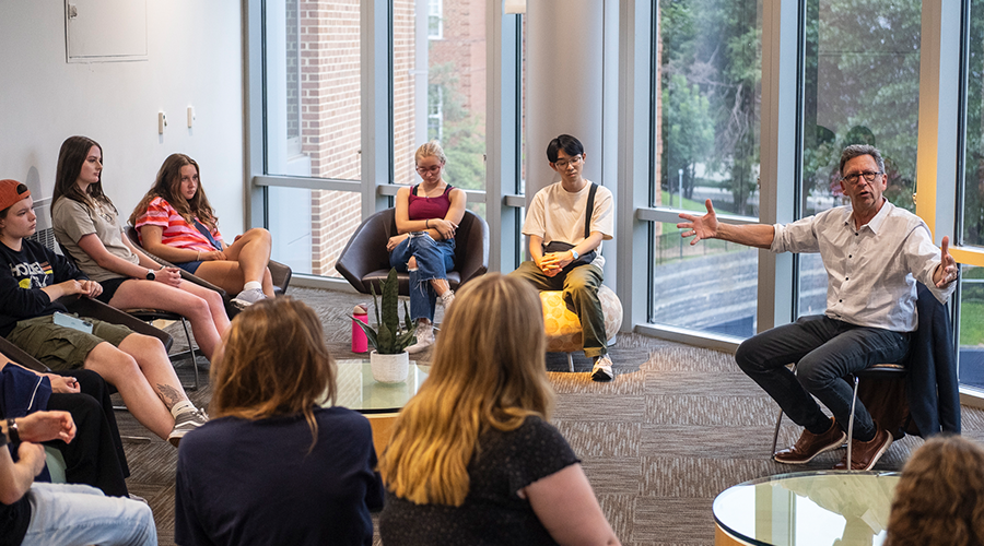 A man in a white collared shirt speaks to a group of students in a room with floor to ceiling windows