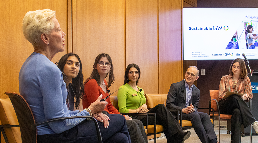 a woman in a light blue shirt speaks to a group of five people on a panel in a room with light wood walls 
