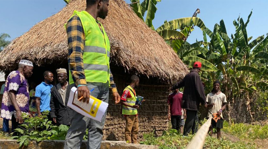 a man in a yellow hi-vis vest stands with a work crew in front of a hut surrounded by grass and trees