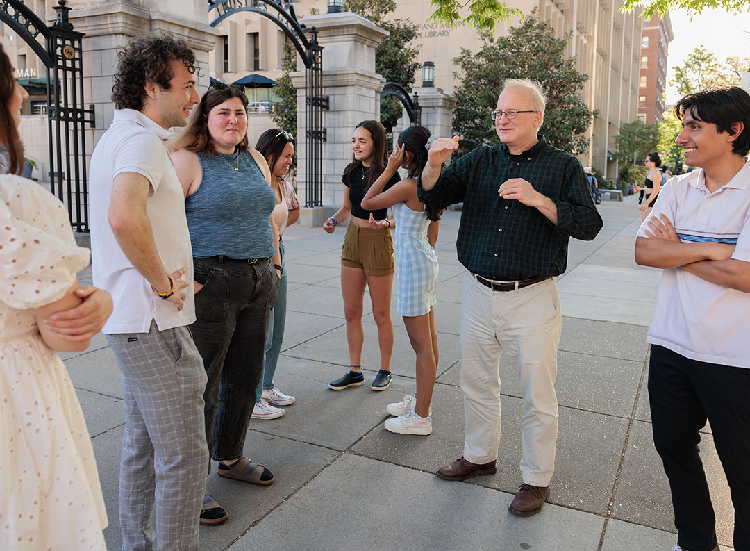 a man in a black shirt talks to a group of seven students on a college campus