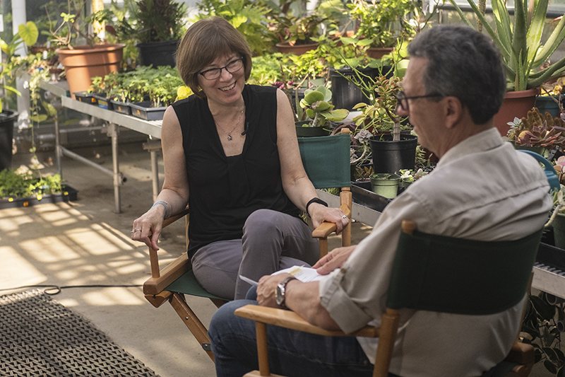 a woman in a black tank top speaks with a man in a beige shirt in a greenhouse