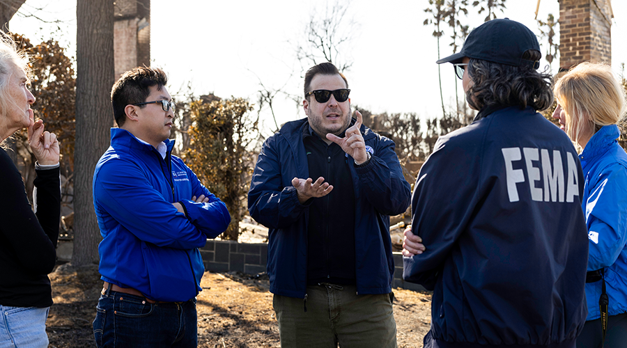 a person in a dark coat speaks and gestures to four people standing outside