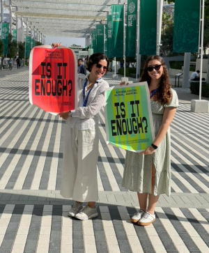 Anna and Hannah stand at the conference holding red and green signs that read "Is It Enough." Anna is wearing a white linen pant and shirt and dark sunglasses and Hannah wears a white and green floral dress. They both have on white sneakers and blue lanyards around their necks.
