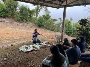 One of the trip leaders leading a teaching session to the GW students, in front of him sits four bags with plants on them, the whole group sits under an awning.