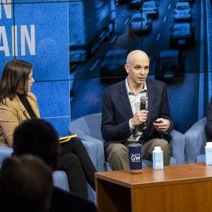 A man in a blue sport coat with a microphone speaks in behind a wood desk in front of a blue background