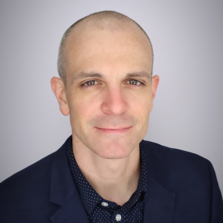 A bald man (JP Halveston) wearing a blue collared shirt and jacket in front of a gray background
