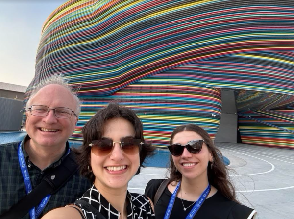 Bob Orttung, Anna Shah, and Hannah Silber in Dubai, UAE. Bob has white hair and glasses and is wearing a collared shirt with a blue lanyard. Anna has short pixie cut brown hair and is wearing dark sunglasses and a black and white sleeveless blouse with a blue lanyard. Hannah has long brown hair and is wearing a black blouse and blue lanyard. They are standing in front of a striped, colorful exhibit backdrop.