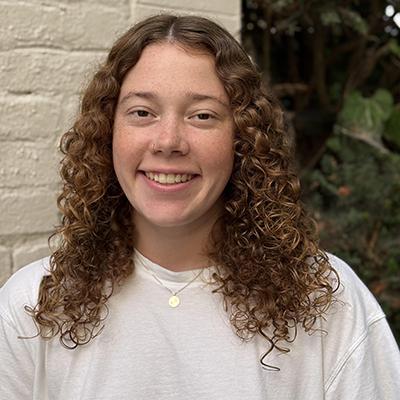 A student with long curly hair in a white shirt next to a wall and foliage