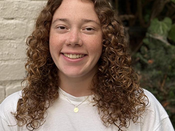 A student with long curly hair in a white shirt next to a wall and foliage