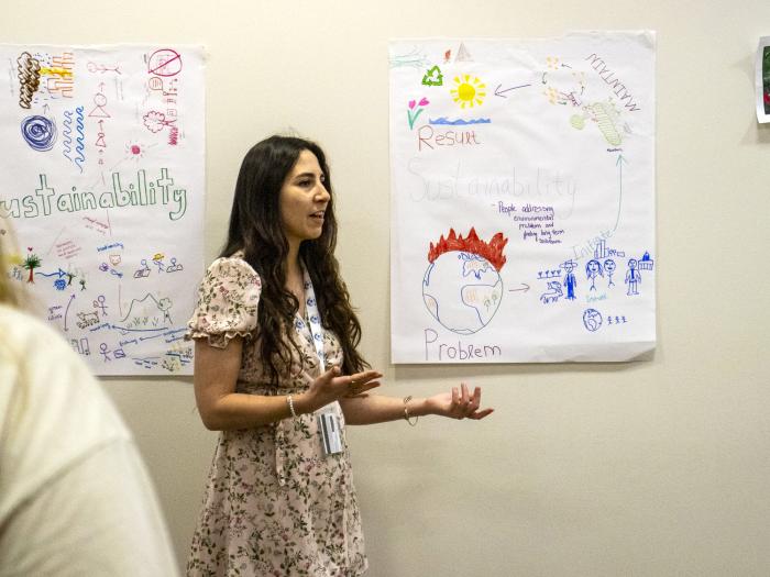 A person with long dark hair stands in front of a poster in a beige room