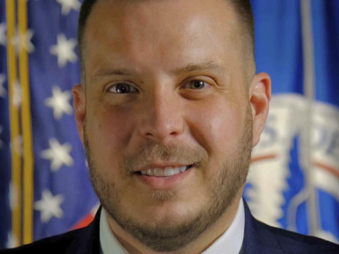a person with a dark suit in front of American and other flags