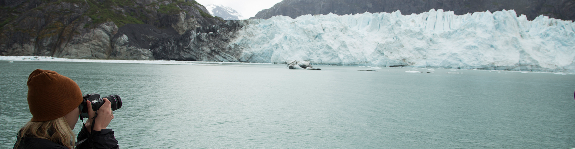 a person in a red knit cap takes a photo of a blue lake and white glacier