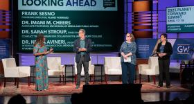 Four people on stage with white chairs behind them, a woman in a colorful dress, a man in a navy sport coat, a woman in a blue shirt, and a woman in a black suit